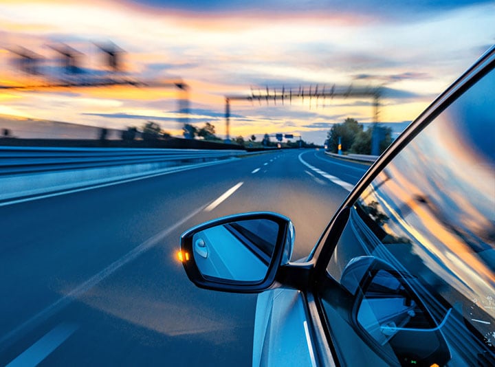 Close up of a car rearview mirror as it drives down the road at sunset.