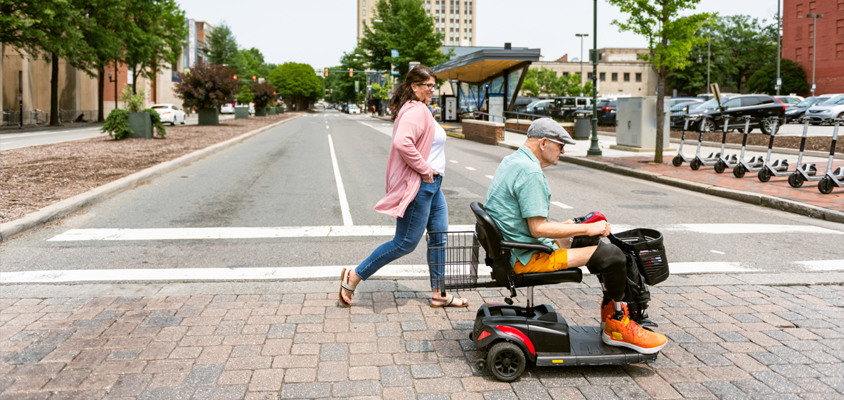 Elderly man on a mobility scooter crossing the street next to a woman walking.